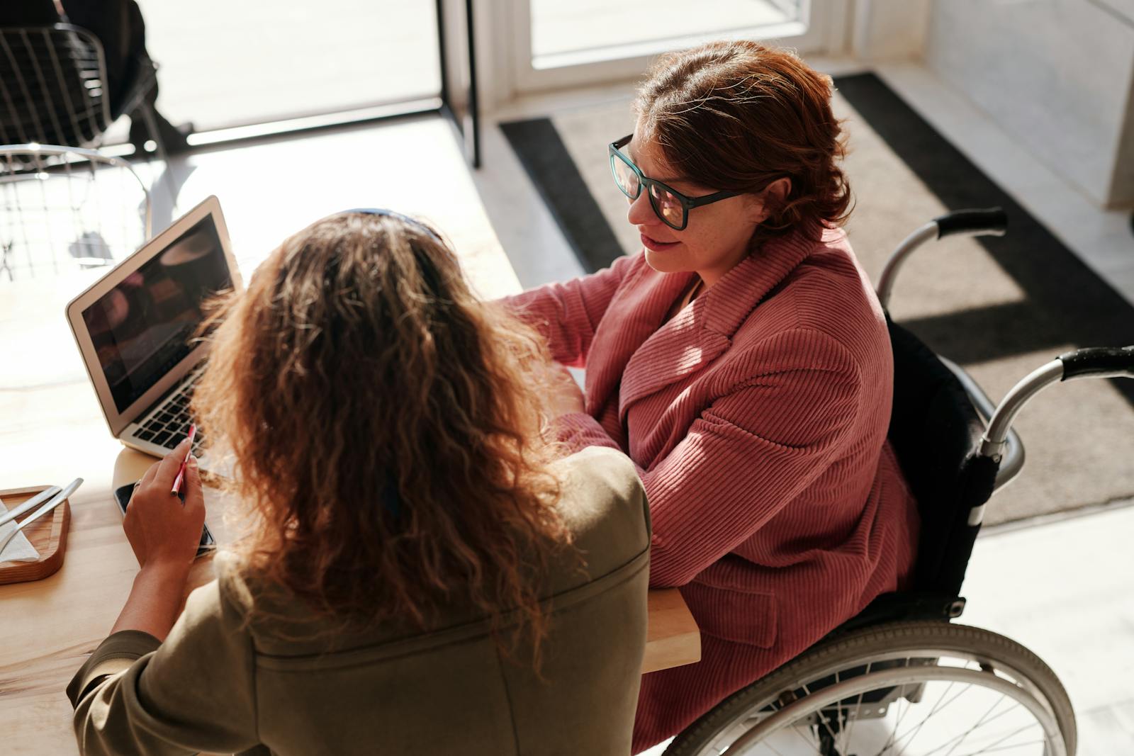 Two women working on laptops, one using a wheelchair, collaborating in a modern café, disability